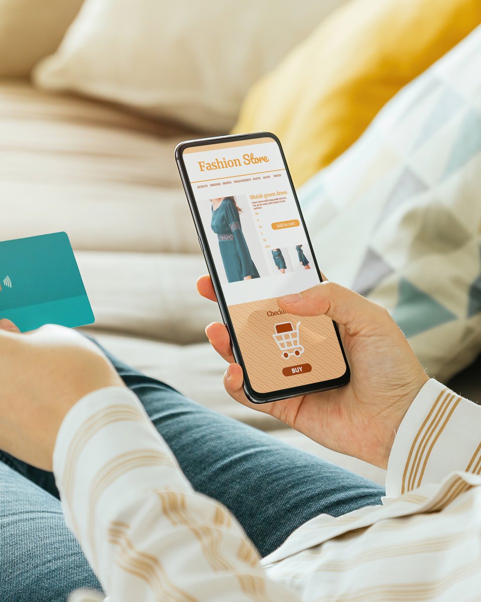 Pessoa deitada no sofá segurando um cartão e comprando roupas em um app de loja no celular.