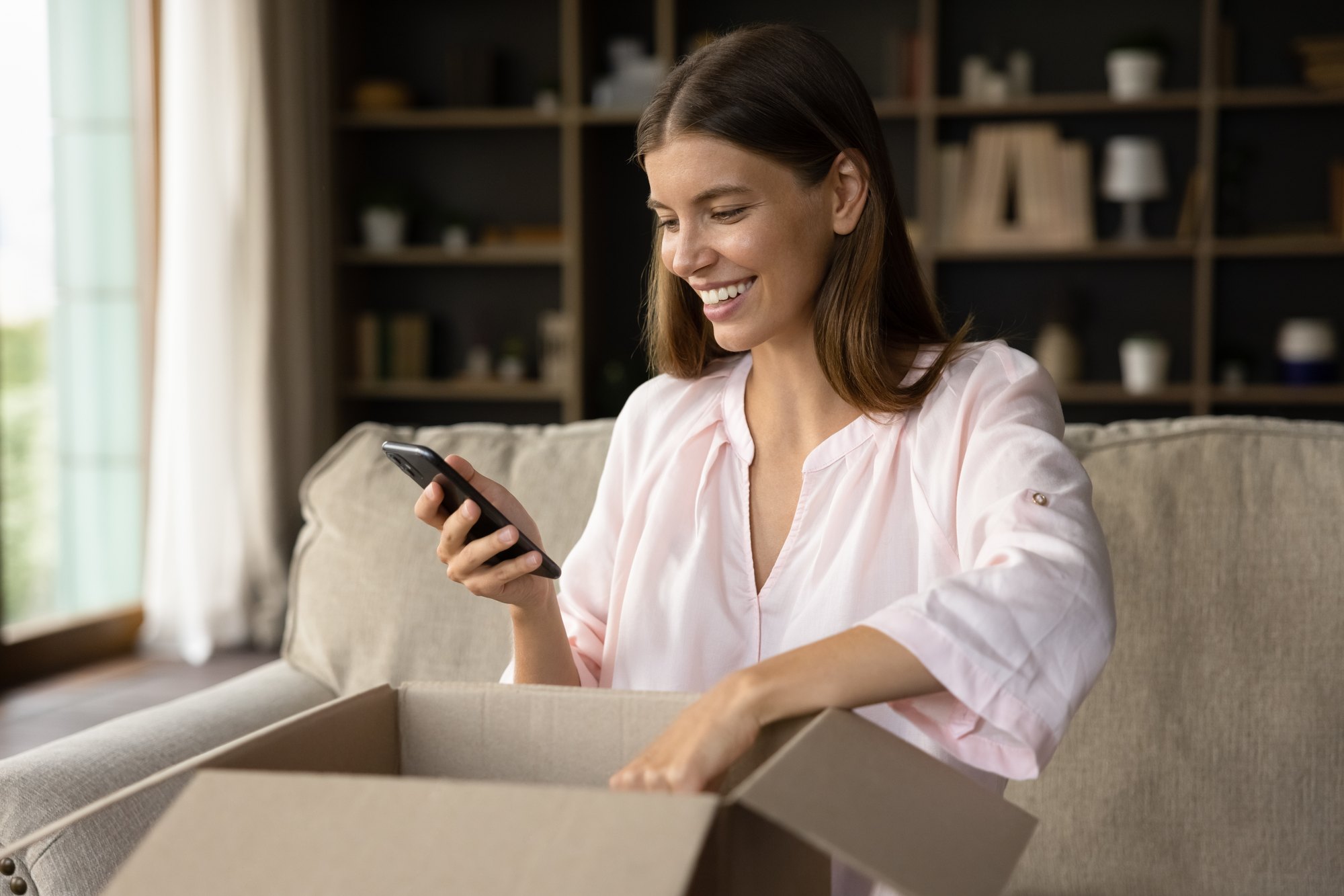 Mulher sorrindo no sofá, olhando o celular enquanto abre uma caixa de papelão de compra online.